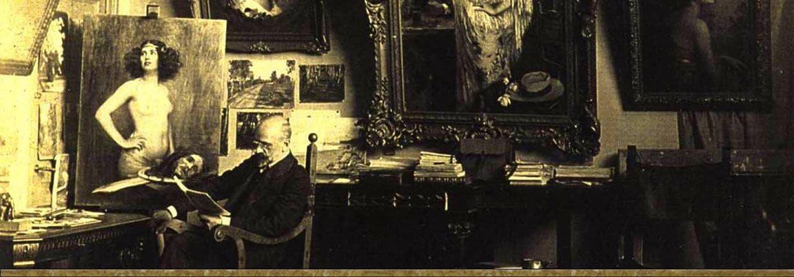 Victor Stember reading in an armchair in his studio, with a painting of a standing nude on the easel and framed works on the wall