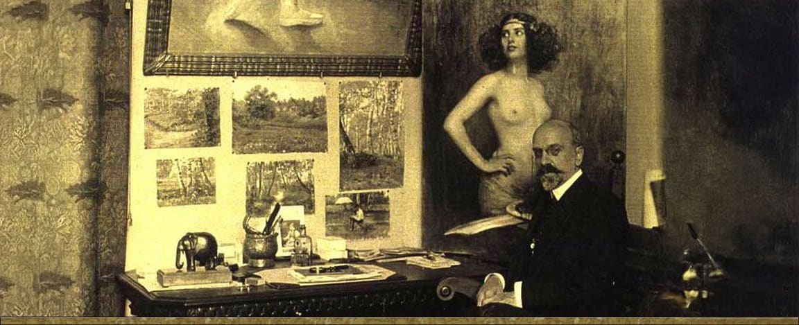 Victor Stember seated at his writing desk, landscape studies pinned to the wall and a painting of a standing nude behind him
