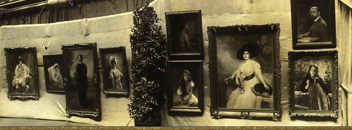 A wall of framed Stember portraits arranged against draped cloth in an exhibition hall, with a potted laurel tree between them