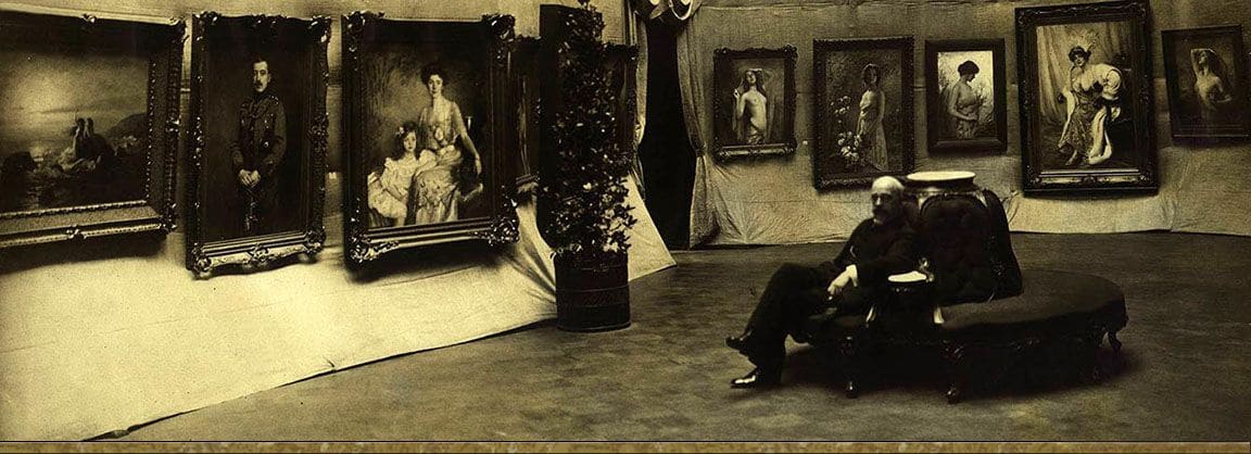 Victor Stember seated on a settee in an exhibition hall, surrounded by his framed portraits hung against draped cloth