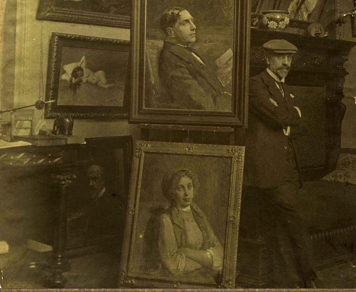 Victor Stember standing with folded arms in his studio, surrounded by framed portraits leaning against the walls and a marble fireplace