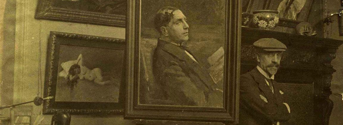 Victor Stember standing with folded arms in his studio beside a portrait of a young man on an easel and a painting of a reclining nude