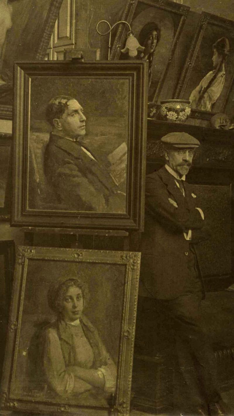 Victor Stember standing with folded arms in his studio beside a portrait of a young man on an easel and a painting of a reclining nude