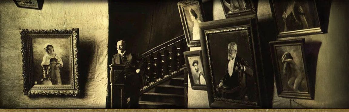 Victor Stember standing at the foot of a wooden staircase among his framed paintings, including a portrait of a girl carrying a jug